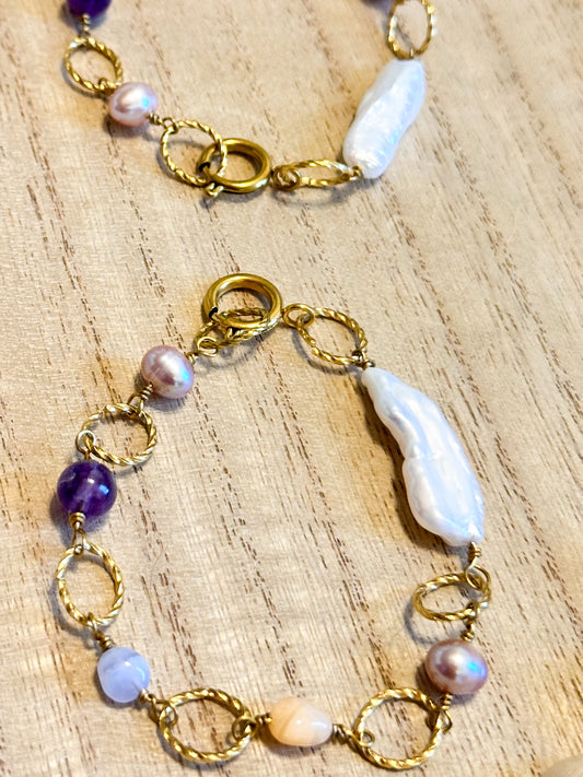 A gold-toned bracelet with twisted gold links and beads, featuring a baroque pearl centerpiece, displayed on a wooden surface.