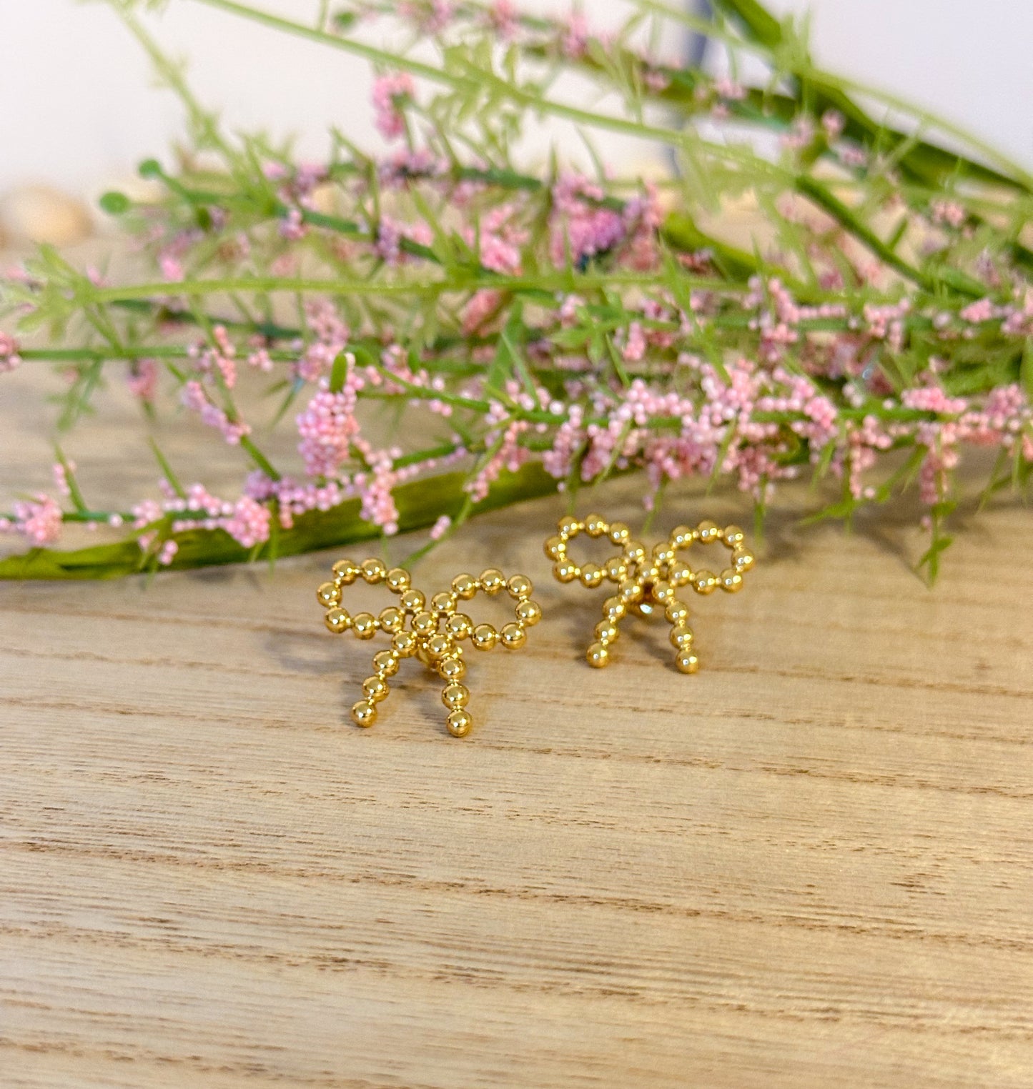 A pair of gold-colored hoop earrings with a dotted bow design, displayed on a wooden surface with a floral background.