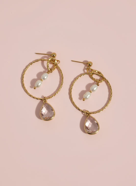 Gold hoop earrings with teardrop-shaped gemstones on a pink background